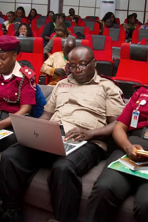 Uniformed agency officials seated among delegates with a laptop and printed materials.