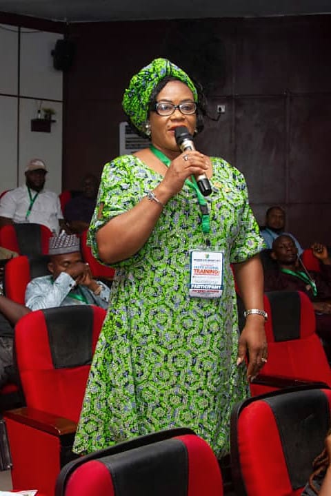 A woman standing with a microphone addressing a seated audience.