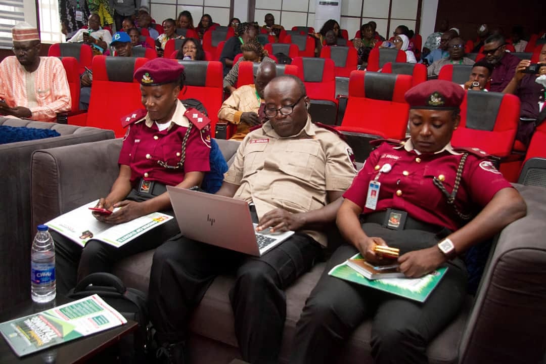 Uniformed agency officials seated among delegates with a laptop and printed materials.